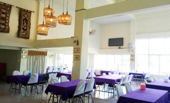 a large dining room with purple tablecloths , white chairs , and hanging lights , under a high ceiling with a television mounted on the at Amonruk  Hotel 2