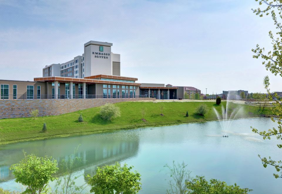 a large hotel with a fountain in front of it , surrounded by grass and trees at Embassy Suites by Hilton Plainfield Indianapolis Airport