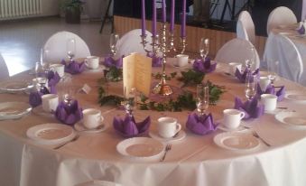 a round table is set with white and purple napkins , wine glasses , candles , and tea cups at Weißes Haus