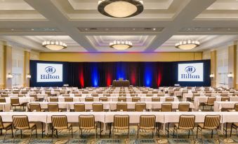 a large conference room with rows of tables and chairs , a stage with two projectors on the wall , and the hilton logo above at Hilton Myrtle Beach Resort