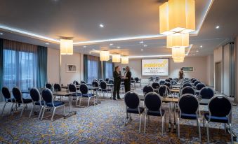 a large , well - lit conference room with rows of chairs arranged in a semicircle , and a projector screen mounted on the wall at Maldron Hotel Belfast International Airport