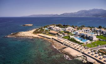 a picturesque beachfront resort with a swimming pool , umbrellas , and lush greenery , situated on a rocky coastline near the water 's edge at Aldemar Knossos Royal