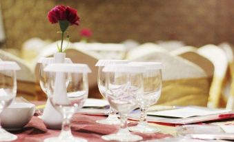 a dining table set with wine glasses , a red flower arrangement , and a book on the table at Sapphire Sky Hotel & Conference