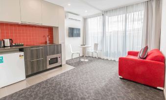 a modern living room with a red couch , gray carpet , and a kitchenette next to a window at City Edge Box Hill Apartment Hotel