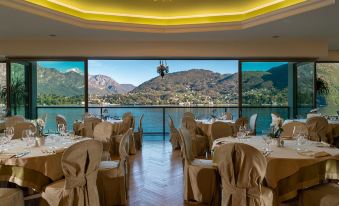 a well - decorated dining room with multiple tables and chairs , surrounded by large windows offering a view of a mountainous landscape at Grand Hotel Cadenabbia