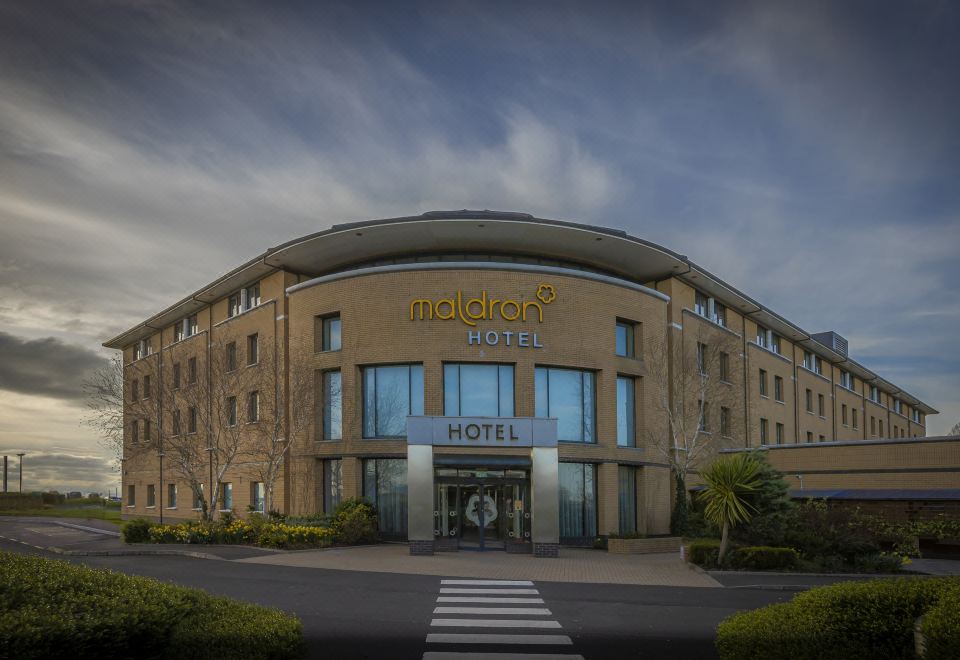 "a large hotel building with a glass facade and the word "" hotel "" displayed on the front" at Maldron Hotel Belfast International Airport