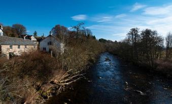 a white house is situated on the side of a river , surrounded by trees and bushes at The Kirkmichael Hotel