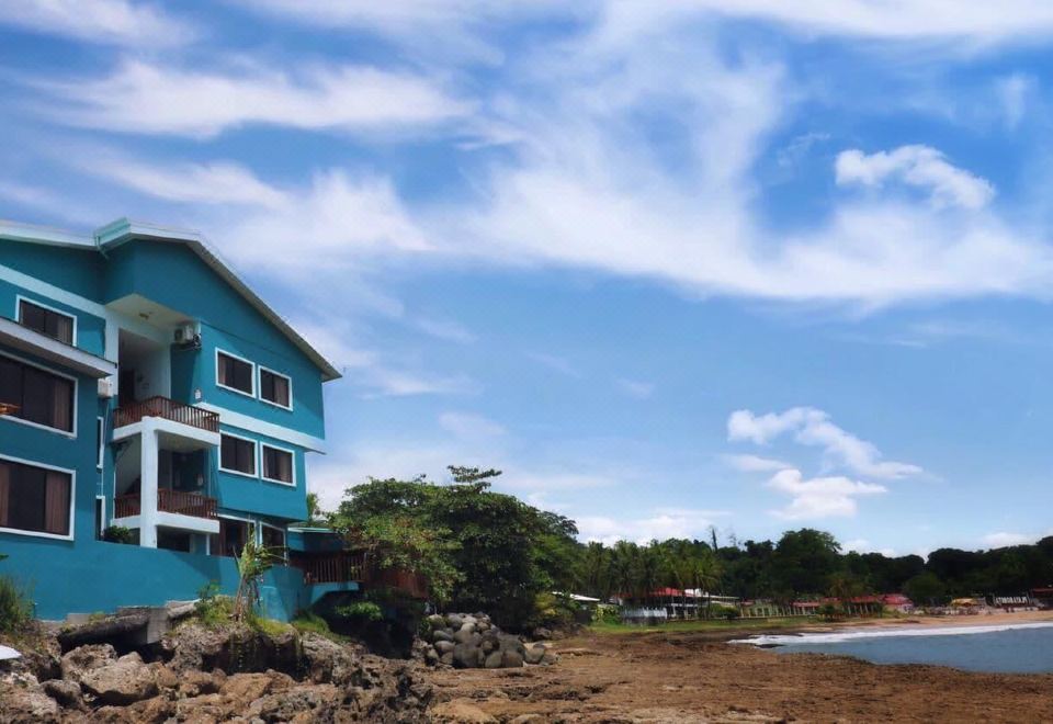 a blue building on the beach , surrounded by rocks and trees , with a cloudy sky in the background at Hotel Playa Bonita