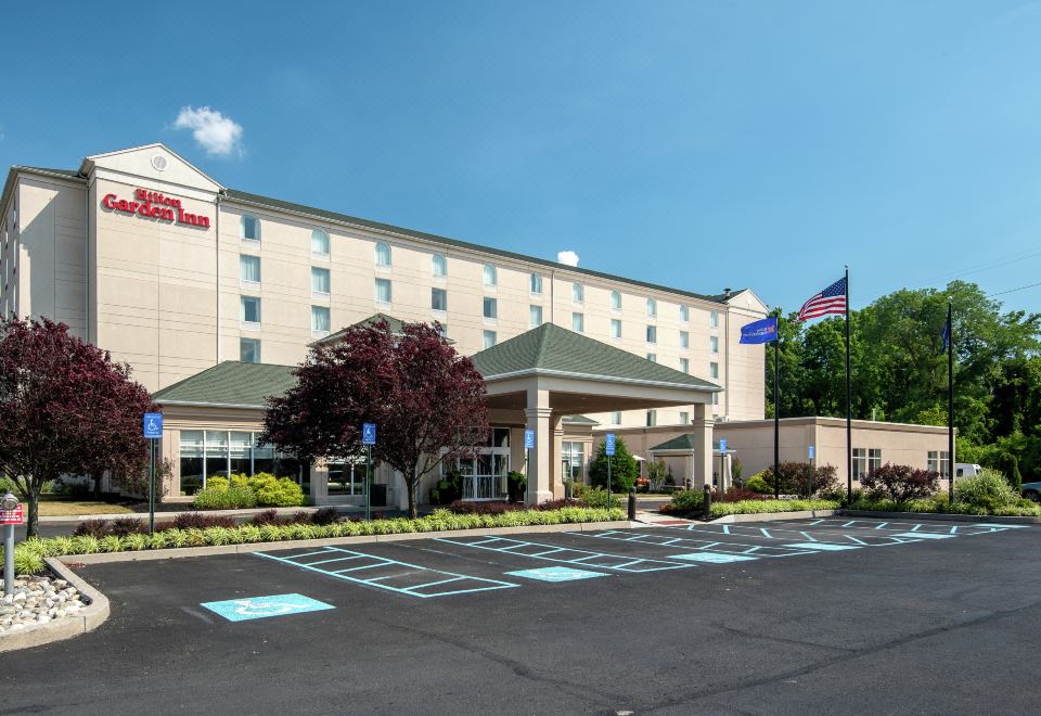 a large hotel with a parking lot in front of it , surrounded by trees and flags at Hilton Garden Inn Philadelphia/Ft. Washington