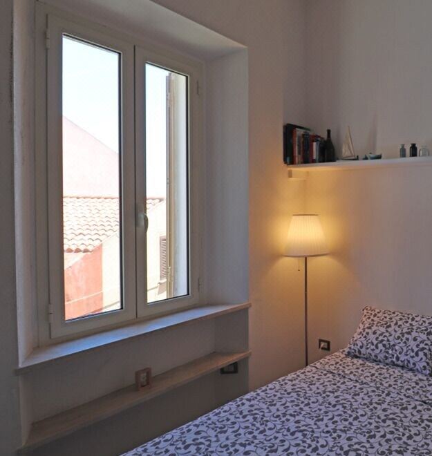 a bedroom with a window that has a lamp on the wall and a book on the shelf at Margherita