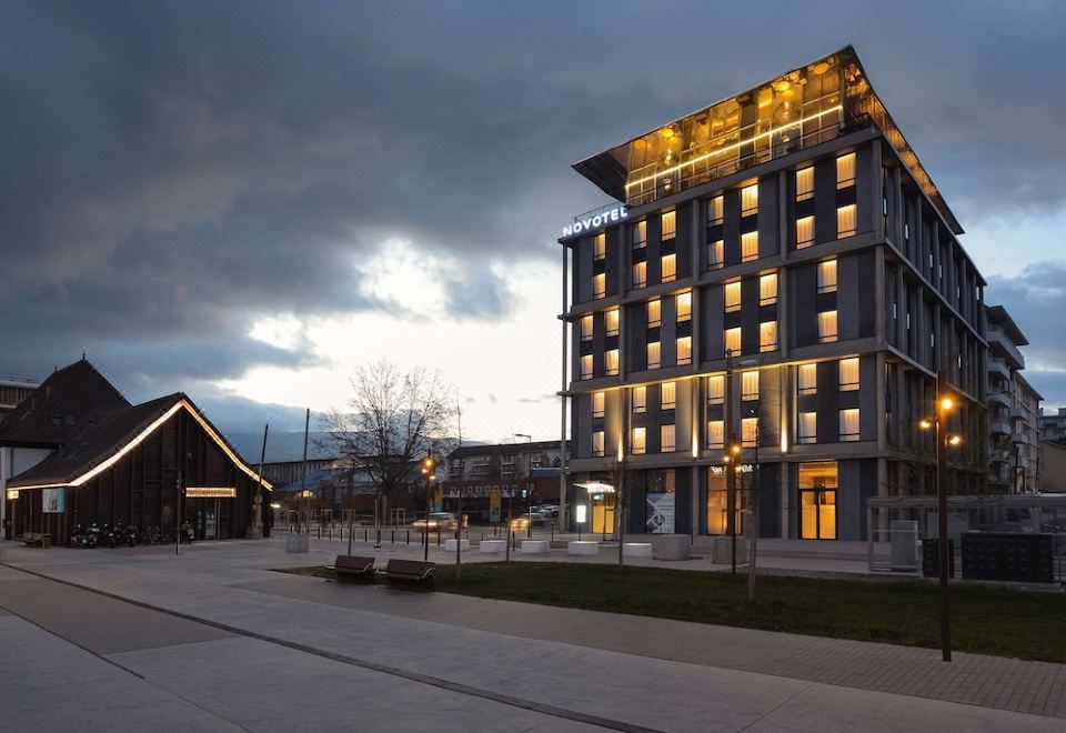 a modern building with a unique design , lit up at night and surrounded by trees and other buildings at Novotel Annemasse Centre - Porte de Genève