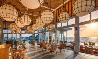 a large , well - lit restaurant with wooden ceilings and wicker furniture , offering a view of the ocean at Raffles Maldives Meradhoo Resort