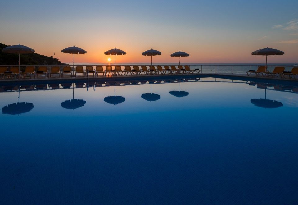 a large outdoor swimming pool surrounded by lounge chairs and umbrellas , with the sun setting in the background at Htop Caleta Palace #HtopBliss
