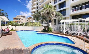 a large swimming pool is surrounded by a pool house and palm trees , with a tall building in the background at Coolum Caprice