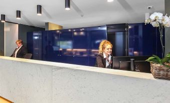 a receptionist is standing behind a white desk in a hotel lobby , smiling and interacting with the receptionist at Belconnen Way Hotel & Serviced Apartments