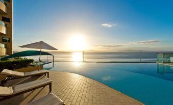 a large swimming pool with a sun umbrella and lounge chairs is seen in the foreground , while the ocean and sky are visible in the background at Majestic Palace Hotel