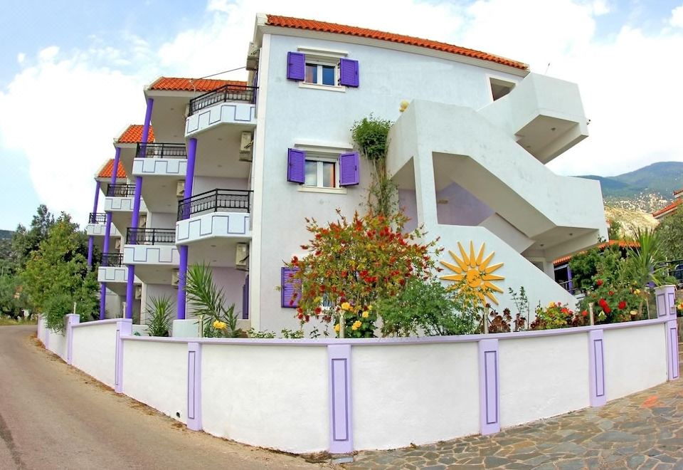 a white apartment building with blue shutters , situated on a street with a flower garden at Lygies Studios