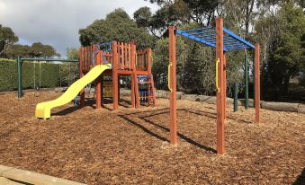 a playground with wooden play structures , including a slide and monkey bars , surrounded by green grass at Prom Coast Holiday Lodge