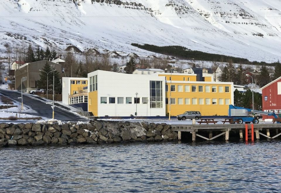 a yellow and white building is situated on the shore of a body of water , with snow - capped mountains in the background at Hildibrand Apartment Hotel