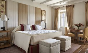 a large bed with white linens and pillows is in a room with beige walls , a window , and a bench at Rosewood Castiglion del Bosco