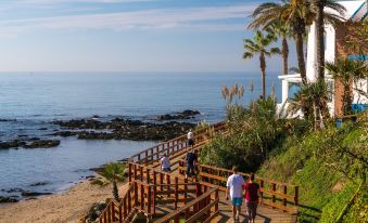 a group of people walking along a wooden boardwalk near the ocean , surrounded by palm trees at El Fuerte Marbella