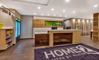 "a modern hotel lobby with a wooden reception desk and a large rug that reads "" home 2 suites by hilton .""." at Home2 Suites by Hilton Battle Creek