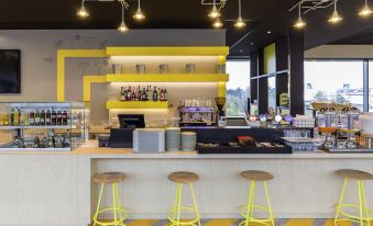 a modern coffee shop with yellow and white furniture , including stools , chairs , and tables , as well as a bar counter at Ibis Styles Geneve Palexpo Aeroport