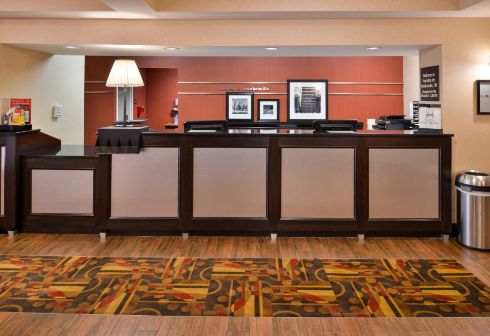 a hotel lobby with a wooden reception desk , a rug on the floor , and several chairs arranged around it at Wingate by Wyndham Steubenville