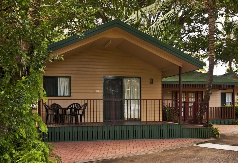 a small wooden house with a covered porch and black chairs on the porch , surrounded by greenery at Cairns Sunland Leisure Park
