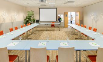 a large conference room with multiple long tables and chairs , a projector screen , and a plant at Ibis Aéroport Bâle-Mulhouse