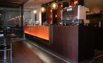 a modern coffee shop with a red and orange counter , hanging lights , and a barista waiting for customers at Hotel Del Corso