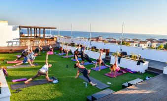 a group of people is doing yoga on a rooftop , with the ocean in the background at E-Hotel Larnaca Resort & Spa