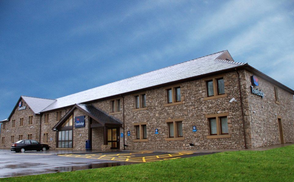 a brick building with a blue sign on the side , situated in a grassy field at Travelodge Kendal