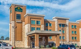a large , tan - colored hotel building with a stone facade and green sign above the entrance , under a clear blue sky at La Quinta Inn & Suites by Wyndham Houston Humble Atascocita