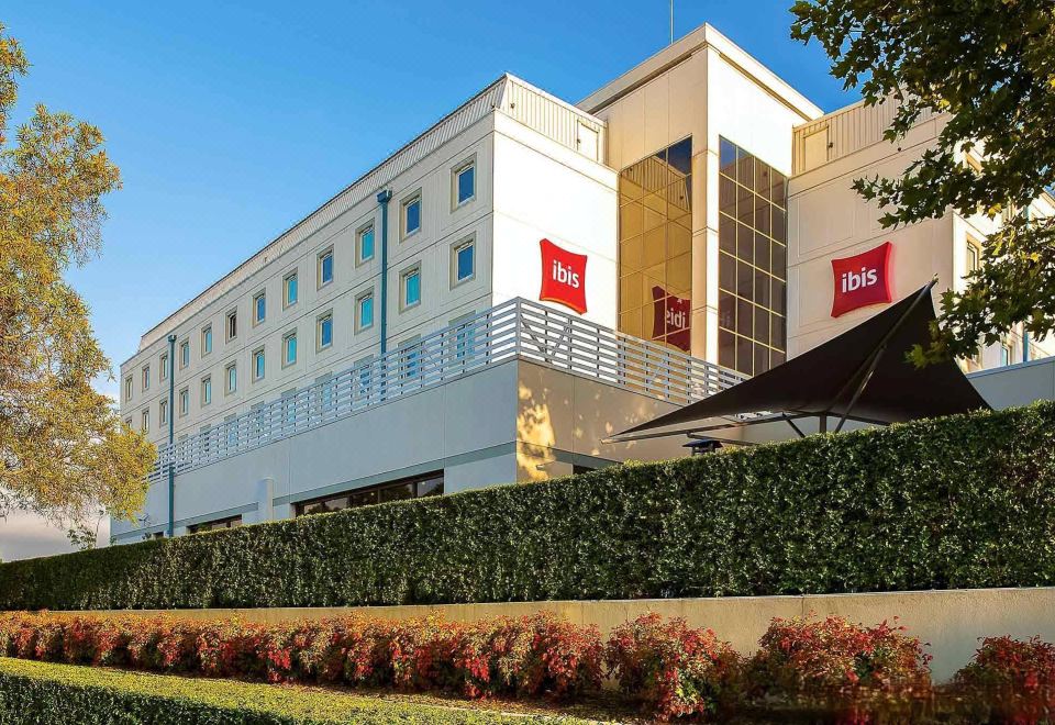 a large hotel with multiple flags on the exterior , surrounded by greenery and a clear blue sky at Ibis Sydney Airport