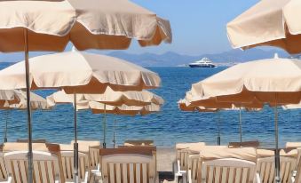 a row of umbrellas with beach chairs on the shore , leading to a sailboat in the water at Cap d'Antibes Beach Hotel