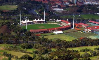 an aerial view of a large , red - roofed stadium surrounded by trees and grass , with two white wind turbines in the foreground at Jacksons Motor Inn