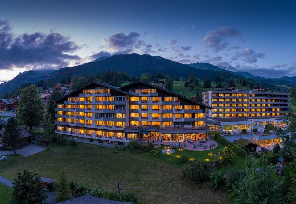 a large hotel building surrounded by grass and trees , with mountains in the background during sunset at Sunstar Hotel & Spa Grindelwald