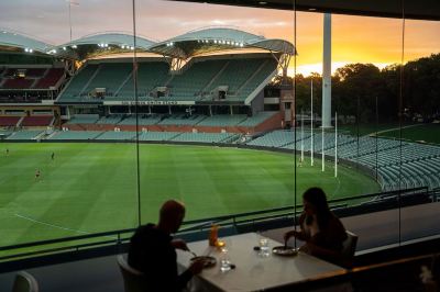 a man and a woman are sitting at a table in front of a large window , overlooking a stadium at Oval Hotel at Adelaide Oval, an EVT hotel