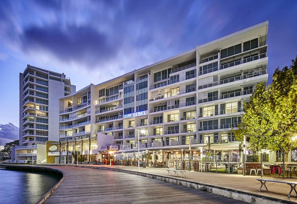 a modern building with multiple floors , balconies , and lights on at night , along with wooden walkways and outdoor seating areas at The Sebel Mandurah