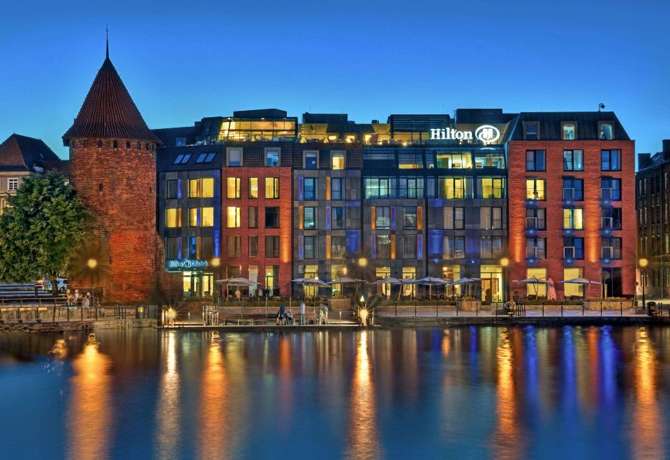 a large hotel building surrounded by water , with the hotel lit up at night , creating a beautiful atmosphere at Hilton Gdansk