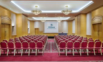 a large conference room with rows of red chairs and a large screen on the wall at Hilton Beirut Metropolitan Palace