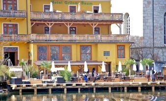 a large yellow building with a restaurant on the side , surrounded by trees and water at Seehotel Grüner Baum