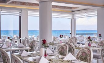 a restaurant with white tablecloths and chairs , set for dining , overlooks the ocean through large windows at Civitel Creta Beach