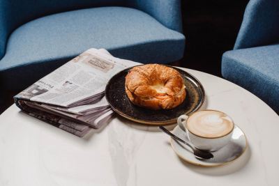 a croissant and a cup of coffee are placed on a table with a newspaper and a chair in the background at Oval Hotel at Adelaide Oval, an EVT hotel