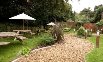 a backyard with a wooden deck , picnic tables , and an umbrella is surrounded by greenery at Premier Inn Burton On Trent East