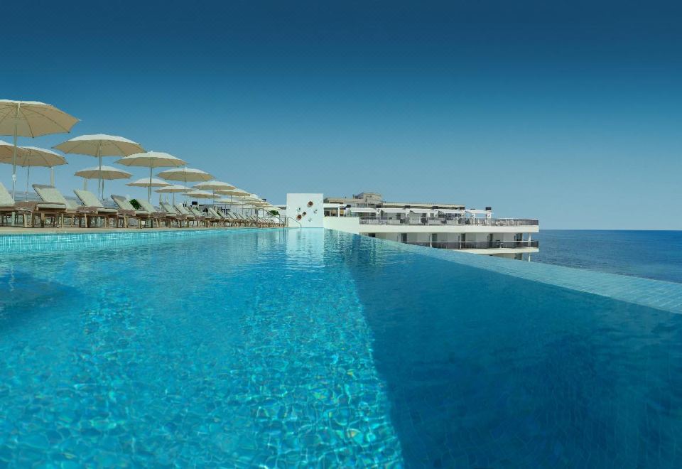 a large swimming pool surrounded by lounge chairs and umbrellas , with the ocean visible in the background at RH Bayren Hotel & Spa