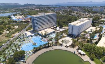 aerial view of a large hotel surrounded by trees and a body of water , with people enjoying their time at the poolside at Club Mac Alcudia