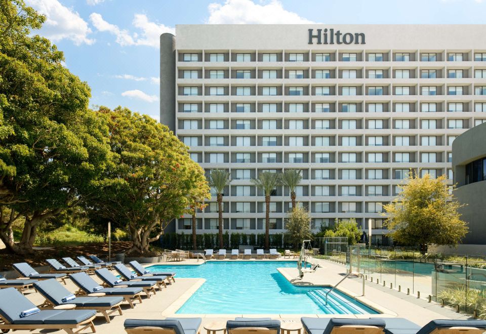 a large hotel with a pool surrounded by lounge chairs and umbrellas , providing a relaxing atmosphere for guests at Hilton Los Angeles-Culver City, CA