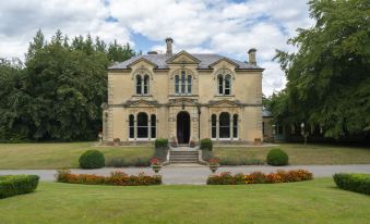 a large , yellow stone building with multiple windows and arched doorways , surrounded by lush greenery and flower beds at Beechfield House
